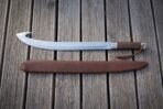 Hooked glaive blade with octagonal wooden grip and braided lanyard laid flat on decking above its brown leather scabbard