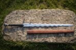 Hooked guard flamberge sword with a polished undulating wave blade, blackened crossguard carrying two parrying hooks, black grip and brown leather scabbard on a rock