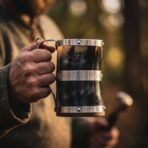 Horn tankard with steel bands, engraved rune band and steel handle held in a man's hand in warm evening light