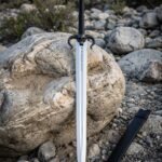 Horned skull sword with a blackened ram horn guard and rune etched blade standing upright on river boulders beside a plain black leather scabbard