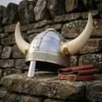 Horned Viking helmet with polished steel dome, brass rivets and knotwork browband resting on a dry stone wall, side angle