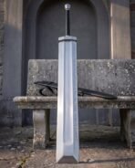 Huge flat steel greatsword with chamfered tip and rune etched blade standing before a stone bench in an arched alcove