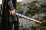 Hunting hanger short sword with polished brass shell guard and knuckle bow held at the side by a cloaked figure on a hillside