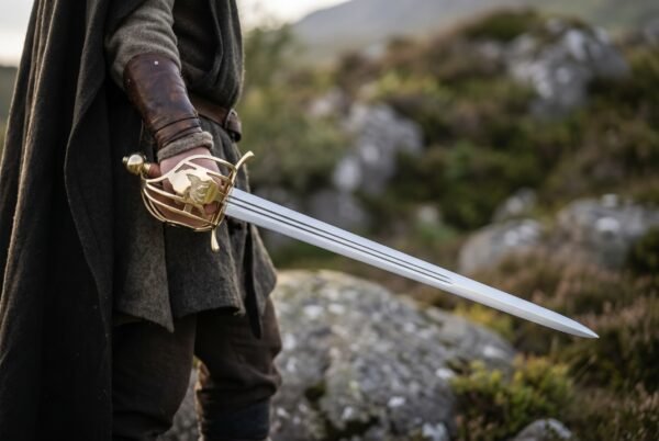 Hunting hanger short sword with polished brass shell guard and knuckle bow held at the side by a cloaked figure on a hillside