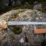 Hunting hanger sword with a polished blade and engraved brass shell guard showing a wolf, resting on a lichen covered boulder beside a tan leather scabbard