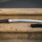 Japanese katana sword with a black and white cord wrapped handle, a brass tsuba and a bright polished blade laid on a sandstone step beside its lacquered saya