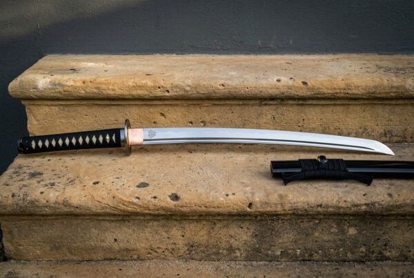 Japanese katana sword with a black and white cord wrapped handle, a brass tsuba and a bright polished blade laid on a sandstone step beside its lacquered saya