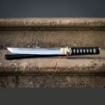 Japanese tanto sword with a mirror polished blade resting on a stone step, showing the engraved brass habaki, round brass tsuba, ray skin handle and black scabbard behind.