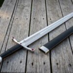 Jewelled arming sword laid beside its black leather scabbard on weathered decking, showing the polished steel disc pommel, red set stones and fullered double edged blade