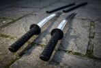 Katana and wakizashi with black cord wrapped tsuka laid side by side on stone paving with their saya in the background