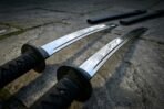 Two katana swords with black cord wrapped handles and round tsuba on grey paving stones, blades curving away from the camera