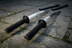 Pair of katana swords with black cord wrapped tsuka laid on old flagstones, two black scabbards resting behind them