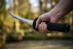 Katana with black cord wrapped tsuka and round black tsuba gripped in both hands, curved steel blade blurred against trees