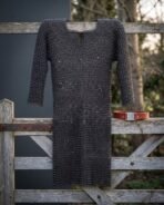 Knee length blackened chainmail hauberk in butted rings hung full length on a wooden gate, red belt with a square buckle plate set beside it
