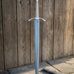 Knightly longsword standing point down against a weathered wooden fence with its pale grey scabbard on the paving, showing the cream cord grip and triquetra shield pommel.