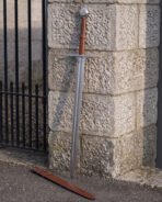 Knightly Norman sword leaning against a granite gate pillar with its russet leather scabbard on the ground, broad fullered blade below a triskele engraved disc pommel