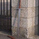Knightly Norman sword leaning against a granite gate pillar with its russet leather scabbard on the ground, broad fullered blade below a triskele engraved disc pommel