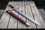 Knightly sword and burgundy scabbard with brass throat and chape laid side by side on a wooden table