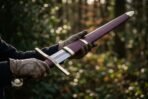 Knightly sword with etched knotwork in the fuller being drawn from its burgundy scabbard by gloved hands