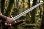 Knightly sword with fullered polished blade and brass cross guard being drawn from an oxblood leather scabbard