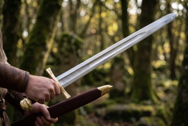 Knightly sword with fullered polished blade and brass cross guard being drawn from an oxblood leather scabbard