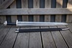 Knotdrift celtic knot panel viking sword laid on grey decking beside its black leather scabbard, showing the pattern welded blade, etched knot band and three lobed steel pommel