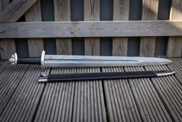 Knotdrift celtic knot panel viking sword laid on grey decking beside its black leather scabbard, showing the pattern welded blade, etched knot band and three lobed steel pommel