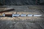Knotwork arming sword with a brown wound leather grip and cut ring chain panel lying on grey paving stones beside a brown leather scabbard in low sun
