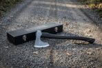 Knotwork bearded axe head in hand forged steel on a dark stained hardwood haft, resting on gravel beside a black engraved wooden presentation case