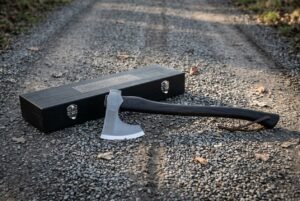 Knotwork bearded axe head in hand forged steel on a dark stained hardwood haft, resting on gravel beside a black engraved wooden presentation case