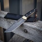 Knotwork damascus bowie resting on weathered grey timber with its black leather sheath behind, showing the etched Celtic panel, brass bolster, black scales and leather lanyard.