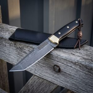 Knotwork damascus bowie resting on weathered grey timber with its black leather sheath behind, showing the etched Celtic panel, brass bolster, black scales and leather lanyard.