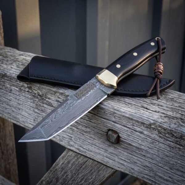 Knotwork damascus bowie resting on weathered grey timber with its black leather sheath behind, showing the etched Celtic panel, brass bolster, black scales and leather lanyard.