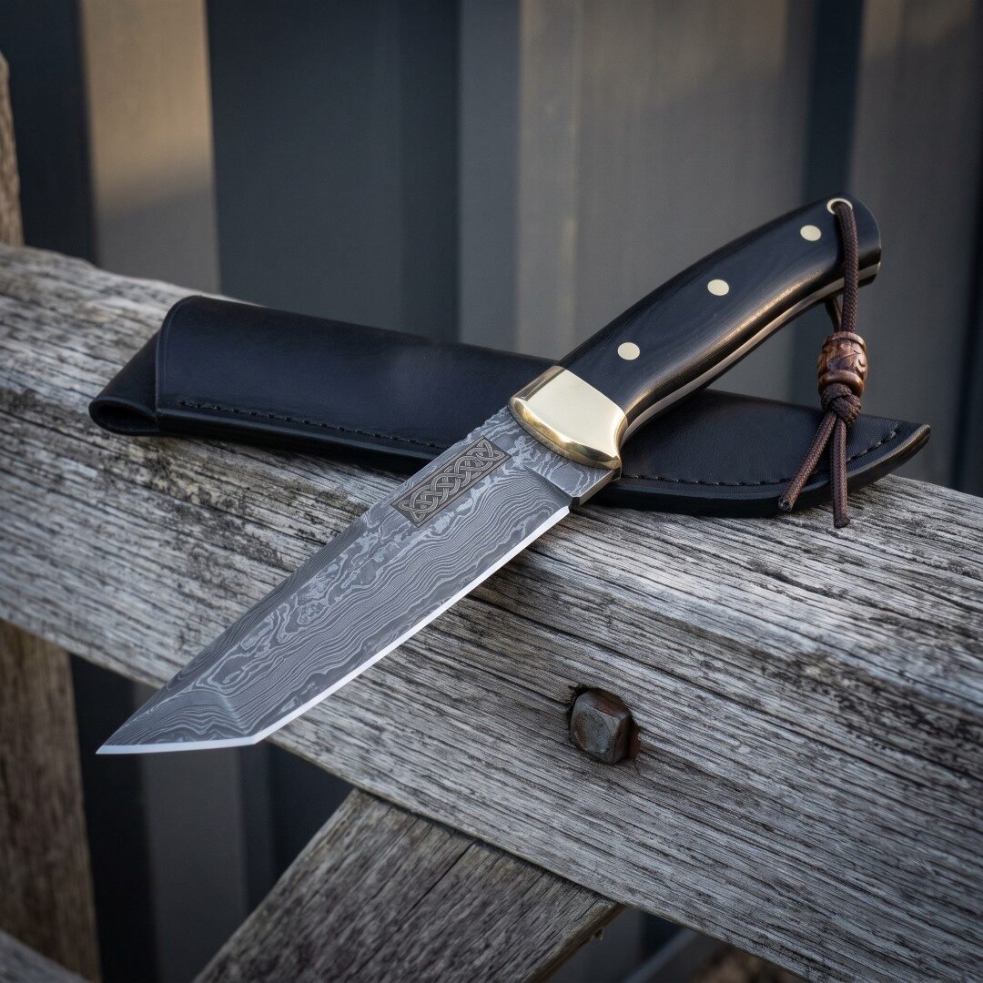 Knotwork damascus bowie resting on weathered grey timber with its black leather sheath behind, showing the etched Celtic Knotwork damascus bowie resting on weathered grey timber with its black leather sheath behind, showing the etched Celtic panel, brass bolster, black scales and leather lanyard.
