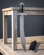 Knotwork damascus falchion standing against a weathered wooden table, its sawback spine, Celtic knot guard and black leather sheath with steel throat clearly shown