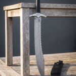 Knotwork damascus falchion standing against a weathered wooden table, its sawback spine, Celtic knot guard and black leather sheath with steel throat clearly shown