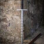Knotwork flamberge sword with a forged Damascus wave blade, cut knotwork guard panel and steel wheel pommel standing against an old sandstone wall beside its scabbard