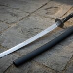 Knotwork katana with a mirror polished 1095 steel blade, etched Norse band, blackened round tsuba and black cord handle lying on grey flagstones beside its saya