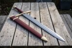 Knotwork knightly sword with a brass wheel pommel and burgundy leather grip lying on a weathered wooden table beside its matching burgundy and brass scabbard