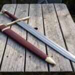 Knotwork knightly sword with a brass wheel pommel and burgundy leather grip lying on a weathered wooden table beside its matching burgundy and brass scabbard