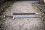 Knotwork panel damascus sword photographed from the side on gravel with its brown scabbard behind, showing the pattern welded flat, etched border band and polished steel chape.