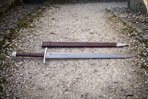 Knotwork panel damascus sword photographed from the side on gravel with its brown scabbard behind, showing the pattern welded flat, etched border band and polished steel chape.