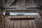 Knotwork viking sword with a five lobed pommel and etched fuller panel leaning against an old barn door beside its dark brown leather scabbard