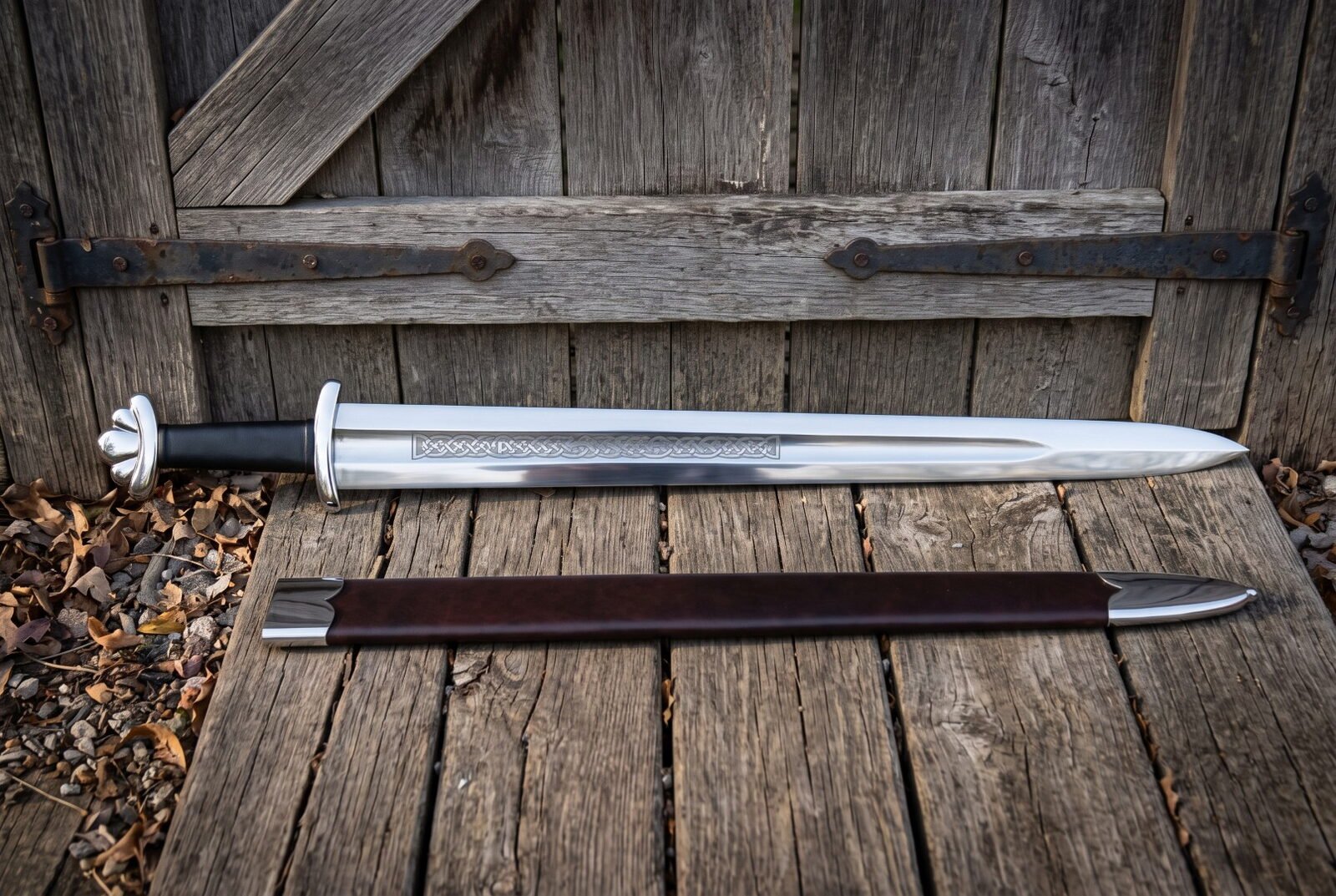 Knotwork viking sword with a five lobed pommel and etched fuller panel leaning against an old barn door beside its dark Knotwork viking sword with a five lobed pommel and etched fuller panel leaning against an old barn door beside its dark brown leather scabbard