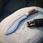 Kukri with stag horn handle and polished curved blade on a rounded stone, companion blades tucked in the leather sheath