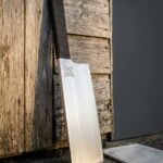 Large hand forged viking cleaver with a Celtic knot engraved on a satin blade and a dark cord wrapped handle leaning against weathered barn boards beside a canvas roll