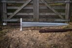 Lattice etched war sword with a broad polished steel blade, brown leather grip and faceted pommel lying on bare earth before a weathered farm gate