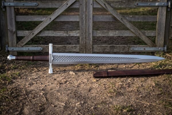 Lattice etched war sword with a broad polished steel blade, brown leather grip and faceted pommel lying on bare earth before a weathered farm gate