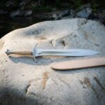 Leaf blade short sword in hand forged 1095 steel with an engraved crossguard and ribbed bone toned grip resting on a pale boulder by a river