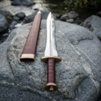 Leaf blade sword with bronze guard and pommel and burgundy leather grip on a boulder beside its leather scabbard